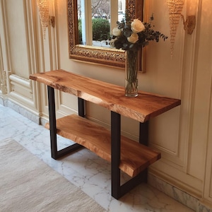 Puede incluir: Una consola de madera con una parte superior de borde natural y un estante inferior. La madera tiene un tono marrón rojizo cálido. La mesa está sostenida por patas de metal negro. Un jarrón de rosas blancas se encuentra sobre la mesa, frente a un espejo dorado.