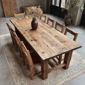 May include: A large, rustic wooden dining table with eight matching chairs. The table is made of light brown, weathered wood and has a rectangular shape. A brown vase with dried flowers sits in the center. The chairs are also made of wood.