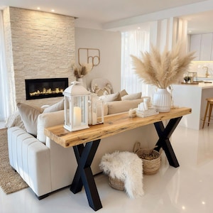 Puede incluir: Un salón con un sofá de color crema, una mesa consola de madera con patas de metal negro y una chimenea. La mesa está decorada con velas, libros y un jarrón con hierba de las pampas. También se ve una linterna blanca y una cesta.