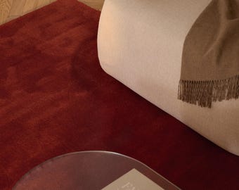 May include: A cream-colored upholstered armchair with a brown fringed throw blanket, positioned on a deep red rug. A round glass coffee table holds a beige book with the text "FA FI" and two small, polished silver-toned metal bowls.