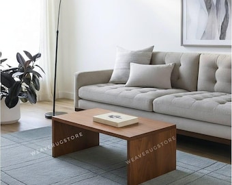 May include: A rectangular, solid wood coffee table with a smooth, medium-brown finish and a waterfall-edge design. The table features the text WEAVERUGSTORE printed in white on its side panels and sits on a large, textured, slate-blue area rug.