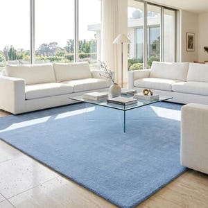 May include: A large, rectangular, solid light blue area rug with a short, dense pile texture. The rug is positioned on a light-colored tiled floor, extending underneath two white sofas and a square glass coffee table in a bright living room.