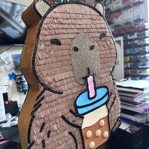 Pode incluir: Uma pinhata marrom em forma de capivara com detalhes brilhantes, segurando um bubble tea. A pinhata tem contornos pretos e um fio branco para pendurar. O copo de bubble tea é azul, branco e marrom com um canudo rosa e branco.