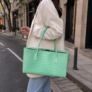 May include: A light green tote bag with a textured crocodile-embossed pattern, featuring two thin shoulder straps and a rectangular silhouette. The bag is carried at the side, showcasing its vibrant color and detailed surface texture.