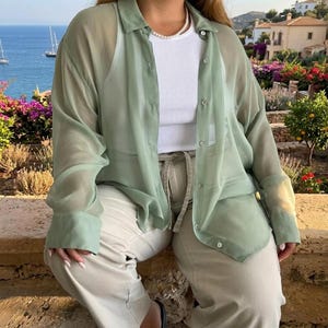 May include: A light green sheer button-down shirt layered over a white ribbed tank top. The outfit includes beige pants and a pearl necklace. The background features a coastal view with boats and buildings.