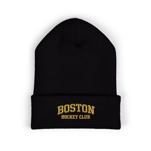 May include: Black beanie hat with gold embroidered text reading "BOSTON HOCKEY CLUB". The hat is a winter accessory, designed to keep the head warm. The text is in a bold, stylized font.