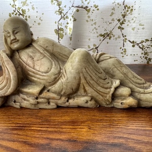 Peut inclure: Une sculpture en pierre beige représentant une figure allongée, peut-être un moine ou une divinité, reposant sur une base texturée. La figure est représentée avec une expression sereine, vêtue de robes drapées. La sculpture est exposée sur une surface en bois.