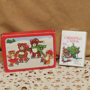May include: A rectangular red box featuring four illustrated brown bears ice skating in a snowy landscape, positioned next to a white deck of playing cards labelled Christmas Cards with a graphic of a decorated tree and a snowman.