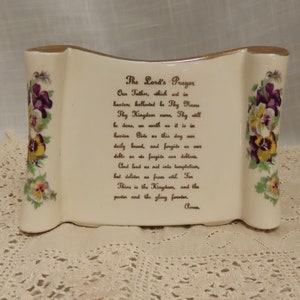 Puede incluir: Un rollo de cerámica blanca con bordes dorados y detalles florales. El rollo presenta la oración del Señor en letras doradas.