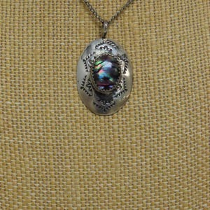 Pode incluir: Um pingente oval de prata com uma incrustação central de concha de abalone iridescente cercada por padrões geométricos estampados. O pingente está suspenso por uma fina corrente de prata, exibindo um acabamento metálico polido com detalhes oxidados escuros.