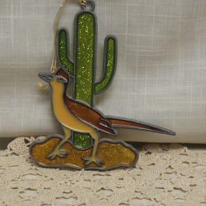 May include: A decorative ornament featuring a roadrunner bird and a green cactus. The roadrunner has brown and yellow coloring, and the cactus has green glitter. The ornament is outlined in a dark gray color and has a gold string for hanging.