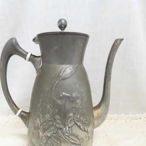 Può includere: Una teiera d'argento con un designo floreale ornato e un coperchio con un pomo a forma di sfera. La teiera ha un beccuccio curvo e un manico.