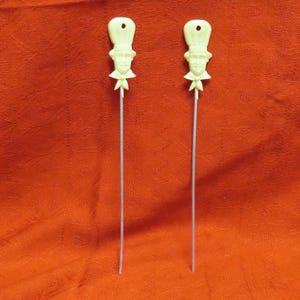 May include: Two off-white chef-themed cocktail picks. Each pick features a chef's face and hat, with a bow tie, atop a thin, silver-colored stem. The picks are set against a textured, red-orange background.