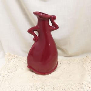 Pode incluir: Um vaso de cerâmica brilhante, na cor bordô, com um design abstrato. O vaso tem um pescoço estreito, um topo alargado e duas alças curvas. O vaso tem aproximadamente 25 cm de altura e está sobre uma toalha de renda.