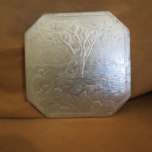 May include: A silver-colored metal trivet with an embossed design of a tree with birds flying around it. There are also figures of people and animals in the design.