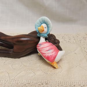 May include: A porcelain figurine of a goose wearing a blue bonnet and a pink dress. The goose is sitting on a wooden surface.