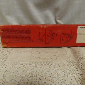 May include: A vintage red box with text and diagrams for a knife sharpener. The box has a white sticker with text. The text on the box includes "COLORADO KNIFE TEETH" and instructions for use.