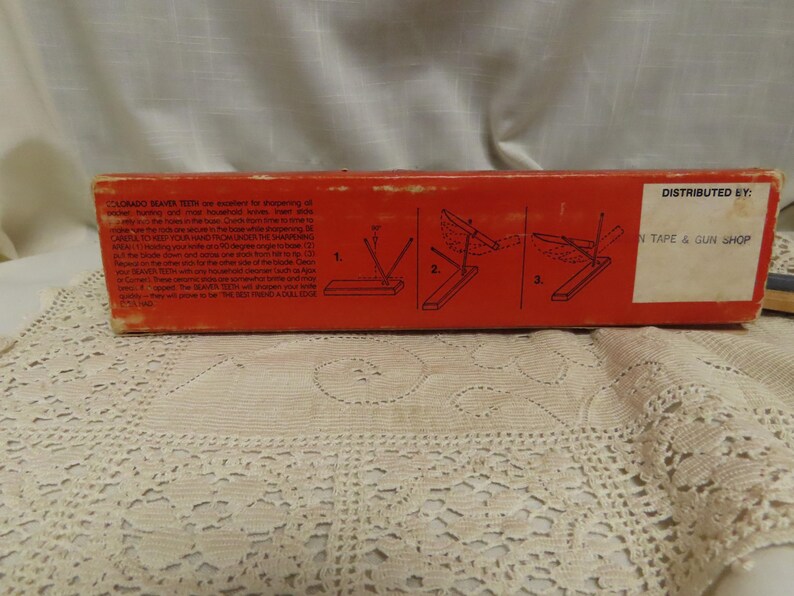 May include: A vintage red box with text and diagrams for a knife sharpener. The box has a white sticker with text. The text on the box includes "COLORADO KNIFE TEETH" and instructions for use.