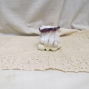 Puede incluir: Un pequeño jarrón de cerámica blanca con base bulbosa y parte superior acampanada. El jarrón tiene un borde morado y detalles dorados. El jarrón está sobre un tapete de encaje color crema.
