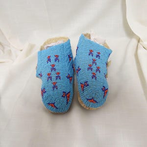 May include: A pair of blue beaded moccasins with red and blue geometric patterns. The moccasins are made of leather and have a fringe detail.
