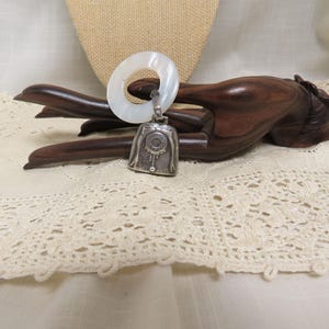May include: A white mother of pearl teething ring with a silver bell charm. The bell has a decorative design and is attached to the ring with a silver loop.