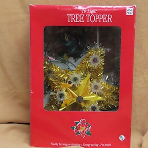 Può includere: Un puntale per albero a 10 luci in una scatola rossa con finestra trasparente. Il puntale a forma di stella presenta orpelli dorati, una stella centrale in lamina d'oro e piccole coperture luminose a forma di stella. Testo: 10 Light TREE TOPPER.