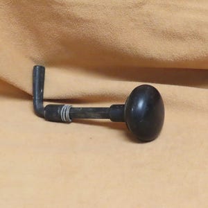 Peut inclure: Une manivelle noire de style vintage avec un bouton arrondi. La poignée a une conception en forme de L pliée et est faite d'un matériau sombre. La poignée repose sur une surface beige.