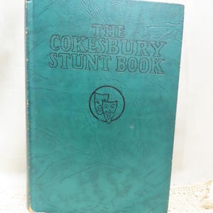 May include: A teal green hardcover book with the title "The Cokesbury Stunt Book" embossed on the front cover. A black and white image of two theatrical masks is centered below the title.