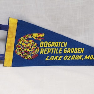 May include: A blue felt pennant with yellow trim and text. The pennant features a coiled snake graphic in yellow, red, and black. Text reads "DOGPATCH REPTILE GARDEN LAKE OZARK, MO."