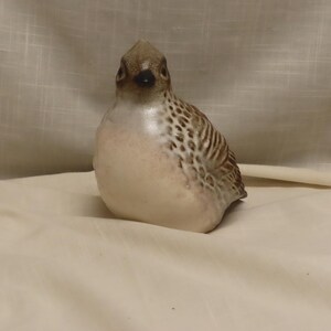 May include: A ceramic bird figurine with a rounded body, featuring a cream-colored chest and a brown-speckled back and wings. The bird has a small, dark beak and detailed markings on its head, presented in a neutral, soft-toned finish.