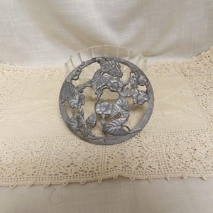 Pode incluir: Um trivet circular de metal prateado com um design floral com folhas e flores. O trivet é colocado sobre uma toalha de mesa de crochê creme e uma tigela de vidro transparente. O fundo é um tecido creme.