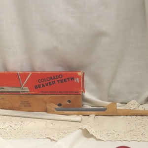 Op de afbeelding: Een antieke gereedschapsset, bestaande uit een houten handvat met een metalen mes, een doos met het opschrift "Colorado Beaver Teeth" en een kleinere doos. De set ligt op een kanten kleedje tegen een witte achtergrond.