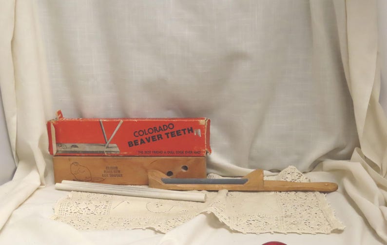 May include: An antique set of tools, including a wooden handle with a metal blade, a box labeled "Colorado Beaver Teeth," and a smaller box. The set is displayed on a lace doily against a white backdrop.