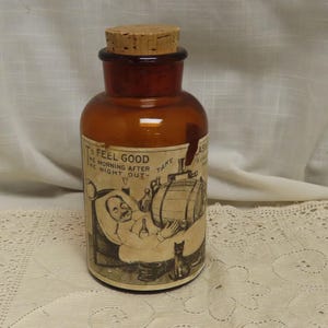 May include: An antique amber glass apothecary bottle with a cork stopper. The label features a humorous illustration and the text "To Feel Good The Morning After The Night Out". A black cat is at the bottom.