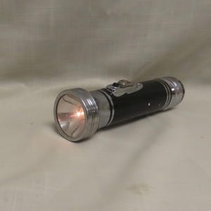 May include: A vintage black flashlight with chrome accents. The flashlight is cylindrical with a silver bezel around the lens and a switch on the side. The light is on, emitting a warm glow. A classic tool for illumination.