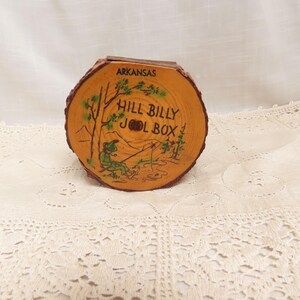 May include: A wooden box shaped like a tree stump with the text "Hill Billy Jol Box" and "Arkansas" on it. The box is decorated with a cartoon of a person fishing.
