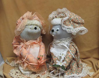 Op de afbeelding: Twee handgemaakte stoffen poppen gemaakt van grijze sokken, gekleed in perzikkleurige en bloemmotief outfits met kanten mutsjes. De linkerpop draagt een bril met een metalen montuur en beide poppen houden kleine kunstbloemen met witte kanten accenten.