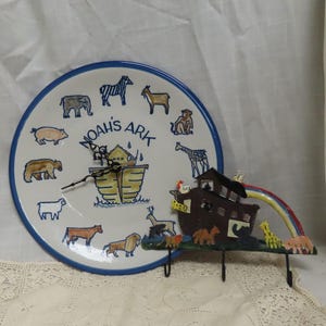 Könnte beinhalten: Ein weißer Keramikteller mit blauem Rand zeigt ein Zifferblatt mit einer Darstellung der Arche Noah und verschiedenen Tieren. Ein Wandhaken aus Metall in Form der Arche Noah mit einem Regenbogen ist ebenfalls abgebildet.