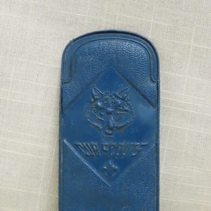 May include: A vintage blue metal case with a diamond-shaped emblem featuring a wolf's head and the words "AIR SCOUTS" above the number 43. The case has a textured surface and a rounded top.