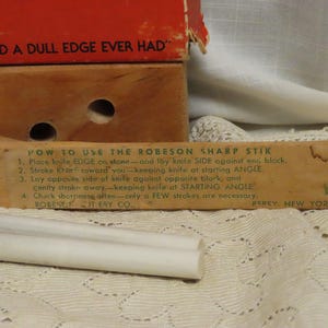 May include: Vintage Robeson Sharp Stik knife sharpener with instructions printed on the wooden handle. The image shows the sharpener, a white sharpening stone, and a red and brown box. The text on the box reads "Best Friend a Dull Edge Ever Had."