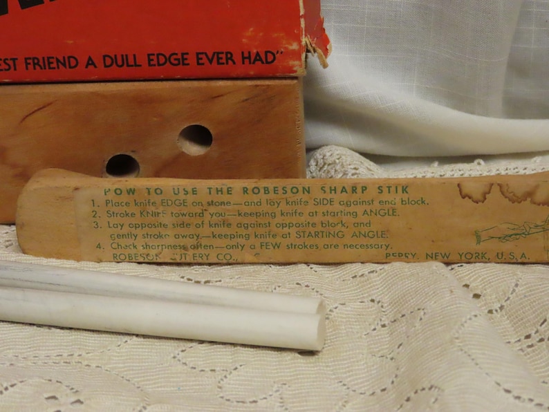 May include: Vintage Robeson Sharp Stik knife sharpener with instructions printed on the wooden handle. The image shows the sharpener, a white sharpening stone, and a red and brown box. The text on the box reads "Best Friend a Dull Edge Ever Had."