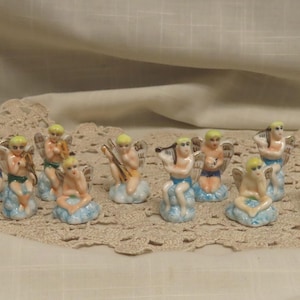 Puede incluir: Una colección de diez pequeñas figuras de ángeles de cerámica, cada una con un tono de piel pálido, alas pintadas de dorado y bases de nubes teñidas de azul. Las figuras se muestran en varias posturas sentadas, algunas sosteniendo instrumentos.