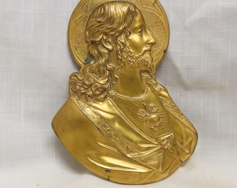 May include: A gold-colored religious plaque depicting a bust of a man with long hair and a beard, surrounded by a halo. The man is wearing a robe with intricate detailing. The plaque has a textured surface and is likely made of metal.