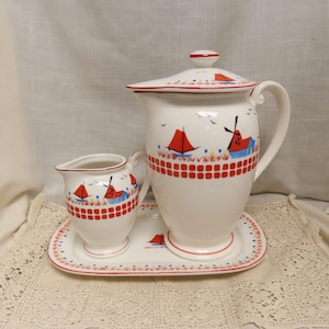 May include: A white ceramic pitcher with a matching creamer and tray. The set features a red and blue nautical design with sailboats and windmills. The pitcher has a lid with a red trim. The set is on a white lace tablecloth.