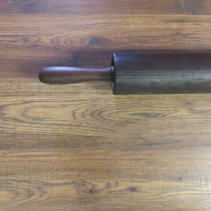 May include: A dark brown wooden rolling pin with a single handle. The rolling pin is cylindrical and appears to be made of a solid piece of wood. The handle is rounded and attached to the main body of the pin.