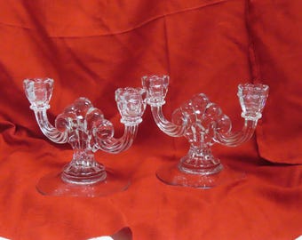 May include: Two clear glass candelabras with a decorative design. Each candelabra has two arms with candle holders and a round base. The candelabras are on a red background.