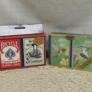 May include: Two vintage playing card decks. One red Bicycle deck with a spade design and the text "Rider Back Playing Cards." The other deck is blue with hummingbird illustrations and the text "Congress Playing Cards." One deck has text that says "Found on the Titanic."
