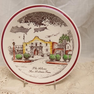 May include: A white ceramic plate with a red rim features a hand-painted illustration of the Alamo in San Antonio, Texas. The plate has a brown and green color scheme and includes the text "The Alamo, San Antonio, Texas."