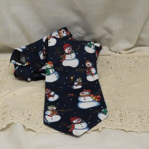 May include: A navy blue necktie featuring a repeating pattern of snowmen. The snowmen have white bodies, black top hats, and colorful scarves and hats. The tie is made of a smooth fabric and is suitable for the holiday season.