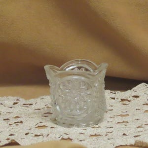 May include: A clear, small, vintage glass toothpick holder with a detailed floral pattern. The holder has a scalloped rim and a decorative design around the sides. The glass is transparent, allowing light to pass through, and it sits on a lace doily.